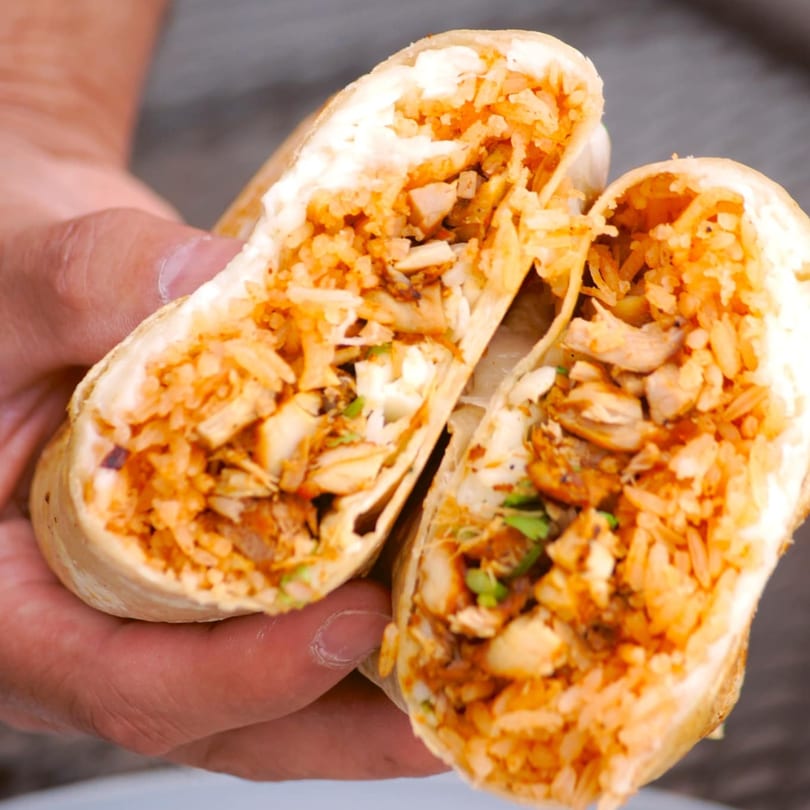 A person's hands holding a large burrito cut in half, revealing chicken, rice, and sauce.