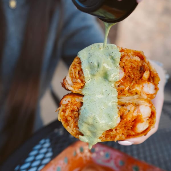 A person holds a large burrito, cut in half, with green sauce drizzled over the top.