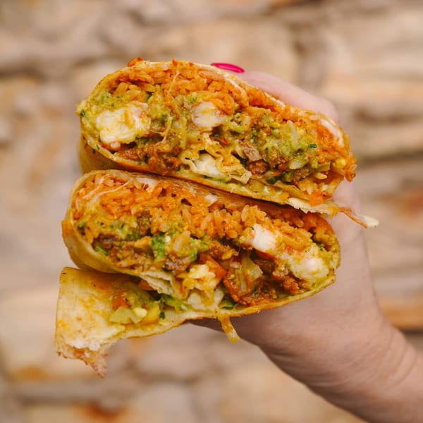 A hand holds a large burrito cut in half, revealing a vibrant filling of rice, meat, and guacamole.
