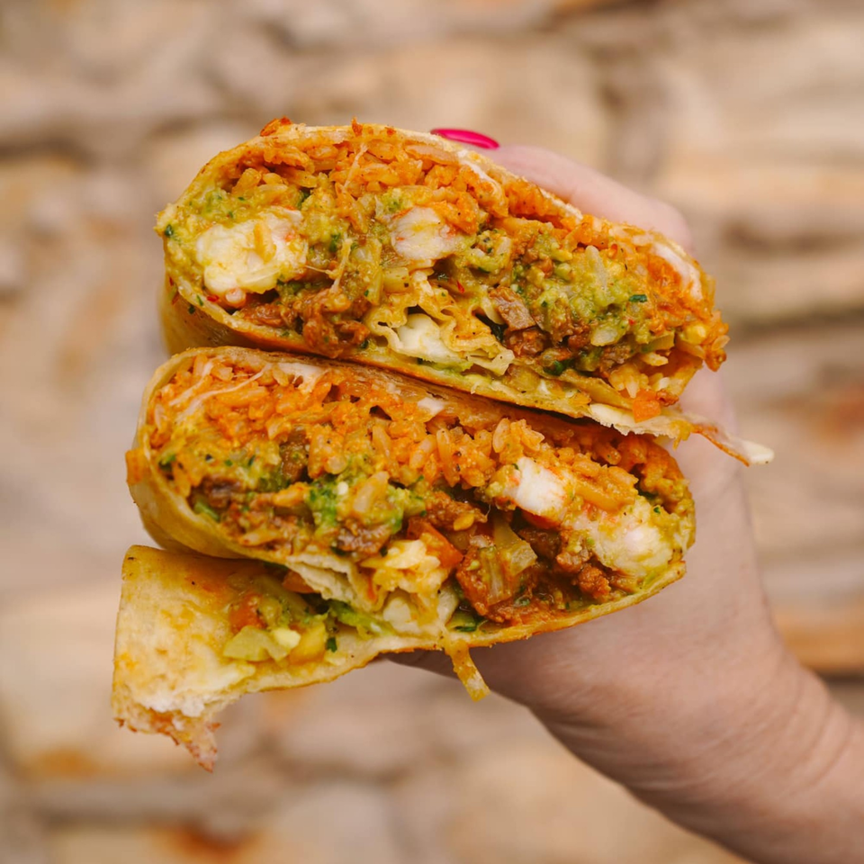 A hand holds a large burrito cut in half, revealing a vibrant filling of rice, meat, and guacamole.